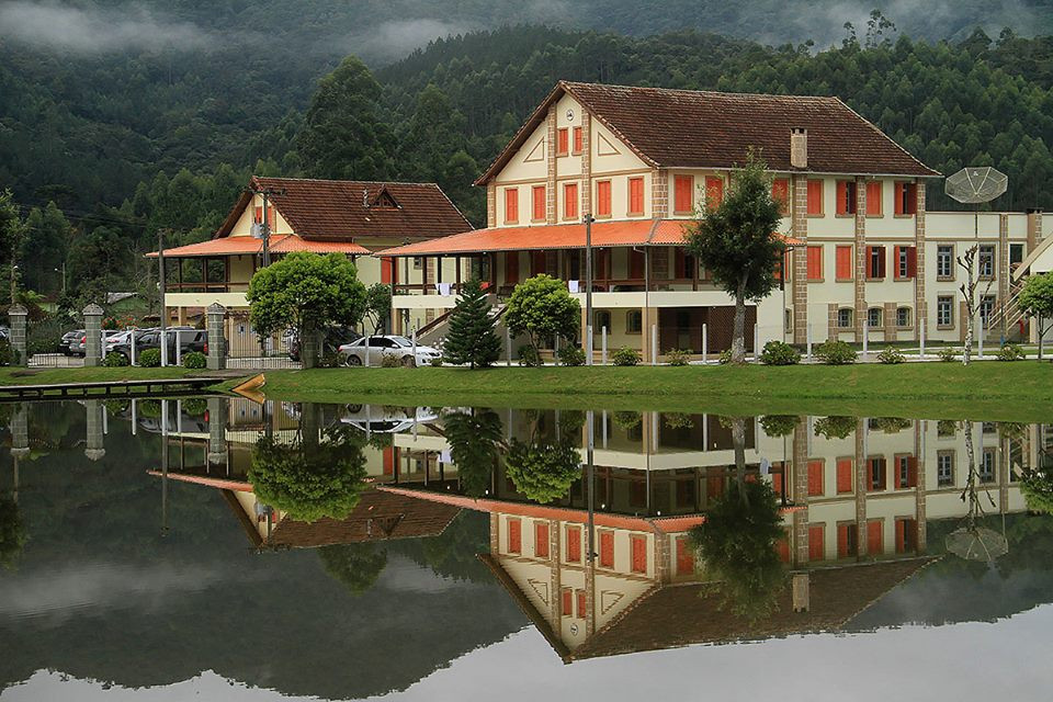 Casarão no centro de Pinheiral refletido no lago, com morros e neblina ao fundo